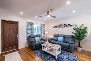 A living room with two sofas, a coffee table, a ceiling fan, a potted plant, and a window with patterned curtains.