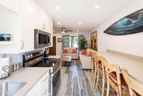 A modern kitchen with white cabinets, a microwave, and a stove. There is a dining area with wooden chairs and a sofa.