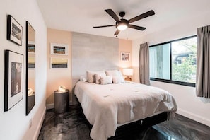 A bedroom with a bed, ceiling fan, artwork on the wall, and a window with curtains.