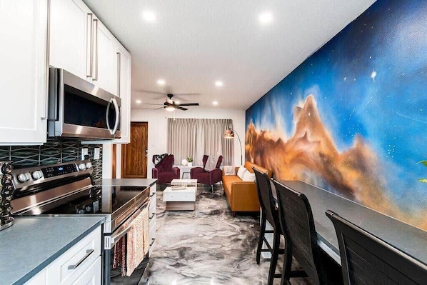 A modern kitchen with a built-in microwave, stove, and oven. There is a dining area with a table and chairs, and a large painting on the wall.