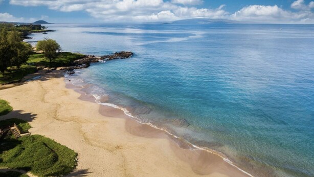 Ein Strand mit klarem, blauem Wasser, sandigem Ufer und etwas Grün.
