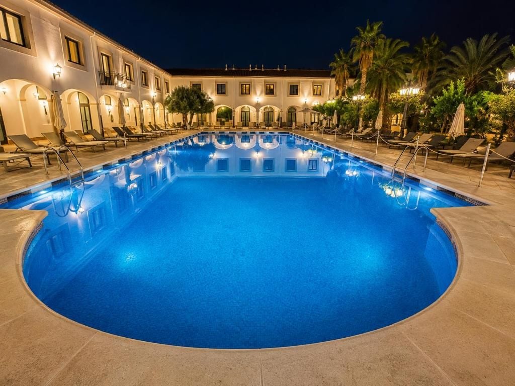 Una piscina con sillones de playa y sombrillas, rodeada por un edificio con ventanas de arco y un balcón.