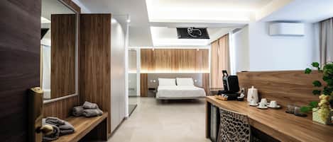 A modern hotel room with a wooden desk, a bed, a bench with towels, and a large mirror.