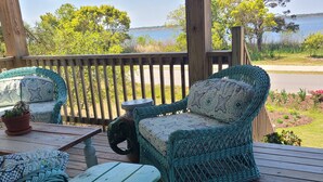 A wooden deck with wicker furniture, including two chairs and a small table, overlooking a body of water. - OBX NEW WATERFRONT DESIGNER HOME WITH DOCK. GORGEOUS AND COMFY INSIDE AND OUT! (Kill Devil Hills)
