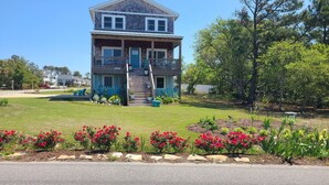 A two-story house with a porch, a staircase, and a well-maintained garden with red flowers. - OBX NEW WATERFRONT DESIGNER HOME WITH DOCK. GORGEOUS AND COMFY INSIDE AND OUT! (Kill Devil Hills)