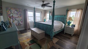 A bedroom with a four-poster bed, a ceiling fan, a bedside table with a lamp, and a colorful rug. - OBX NEW WATERFRONT DESIGNER HOME WITH DOCK. GORGEOUS AND COMFY INSIDE AND OUT! (Kill Devil Hills)