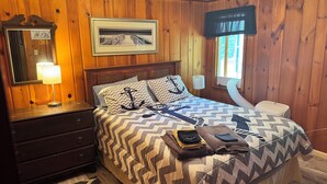 A wooden-paneled room with a bed, a nightstand, a lamp, a framed picture, and a window with a view. - Sailors Delight Back Cottage (Greenbush)
