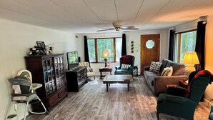 A living room with a wooden floor, a ceiling fan, a television, a sofa, a chair, a coffee table, and a cabinet. - Sailors Delight Back Cottage (Greenbush)