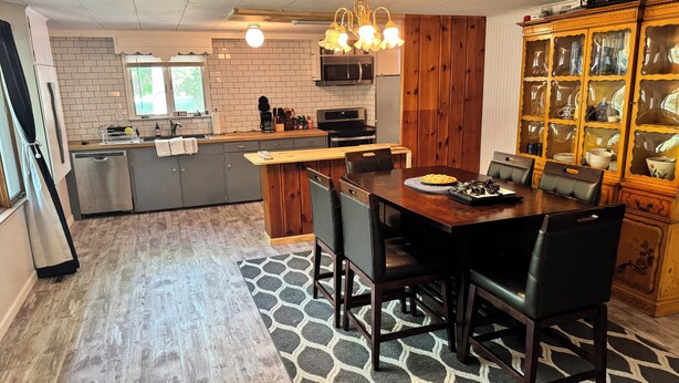 A modern kitchen with a central island, wooden cabinets, and a dining area with a table and chairs. - Sailors Delight Back Cottage (Greenbush)