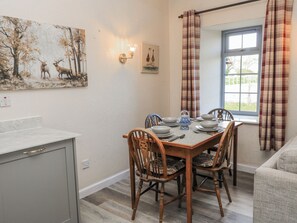 Un comedor con una mesa de madera dispuesta para cuatro personas, sillas alrededor, una lámpara de pared y una ventana con cortinas.