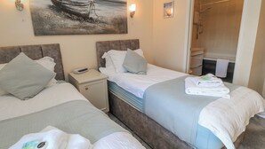 A hotel room with two beds, a nightstand, a painting of a boat on the wall, a bathroom with a shower, and towels on the beds. - Carthouse (Weston-super-Mare)