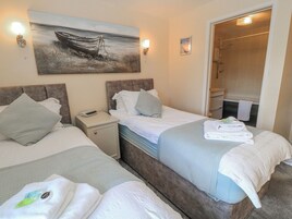 Quarto de hotel com duas camas, uma mesa de cabeceira, um quadro de um barco na parede, banheiro com chuveiro e toalhas sobre as camas.