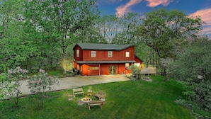 A two-story house with a red exterior, a front porch, and a backyard with a fire pit and seating. - The Red Lodge - Unique Gem Near Lakes with Game Room and Hot Tub! (Elkhorn)