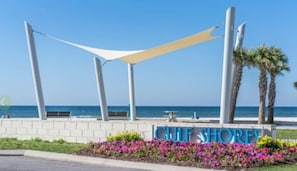 A beachfront sign with 'CLIF SHORE' written in blue letters, surrounded by colorful flowers and a white canopy structure.