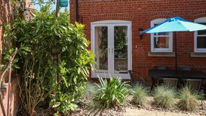 A brick house with a glass door, a patio with a table and chairs, and various plants. - Trinity Cottage (Christchurch)