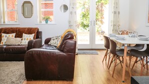 A living room with a brown leather sofa, a dining table with chairs, and a view of a brick wall and greenery outside. - Trinity Cottage (Christchurch)