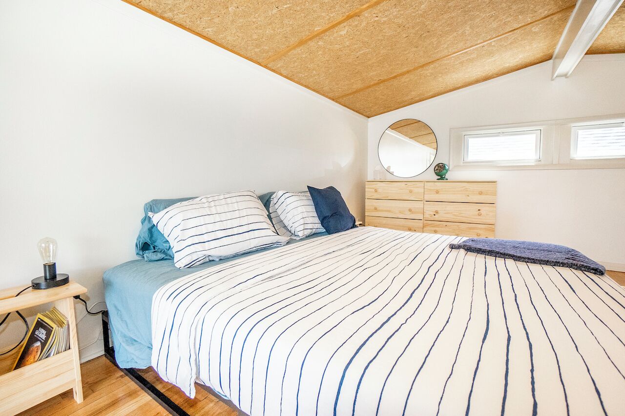 A bedroom with a bed, a wooden dresser, a round mirror, and a small table with a lamp.