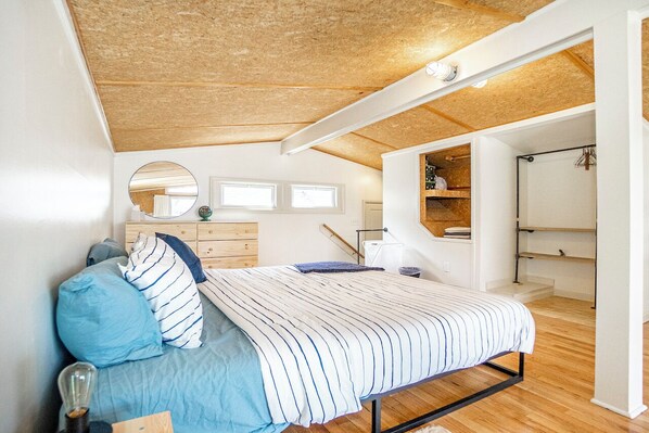 Un dormitorio con techo de madera, una cama con ropa de cama a rayas, un aparador, un espejo y una estantería.