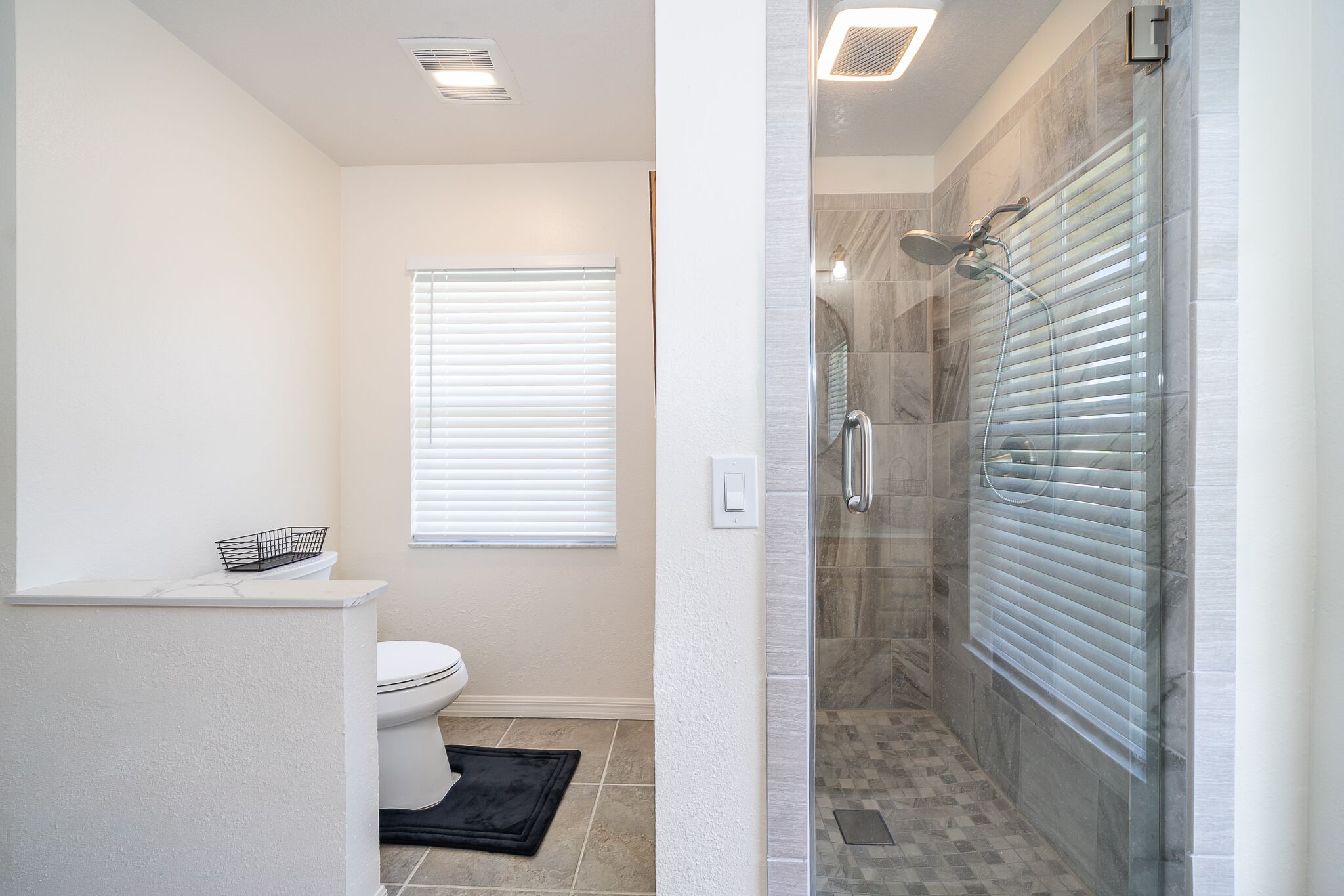 A bathroom with a toilet, a shower with glass doors, and a window with blinds.