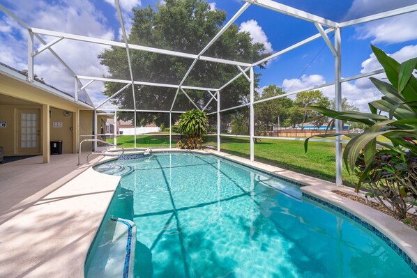 A swimming pool with a clear blue water, surrounded by a white metal structure, and a grassy area with trees in the background.