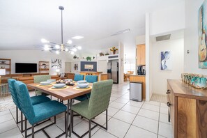 A modern kitchen with a central island, a dining area with teal chairs, and a living room with a flat-screen TV.