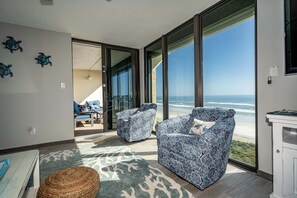 A room with two patterned armchairs facing a large sliding glass door leading to a balcony with a view of the beach. - Sand Dollar 507 Penthouse Paradise (St. Augustine)