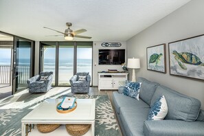 A living room with a view of the ocean, a ceiling fan, a TV, and a sign that says "Welcome to Paradise".