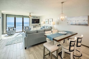 A spacious living area with a large window overlooking the ocean, a grey sofa, a dining table with chairs, and a ceiling fan. - Sand Dollar 507 Penthouse Paradise (St. Augustine)