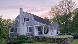 Une maison à deux étages, avec des fondations en pierre, une grande cheminée et une véranda couverte dotée de sièges extérieurs.