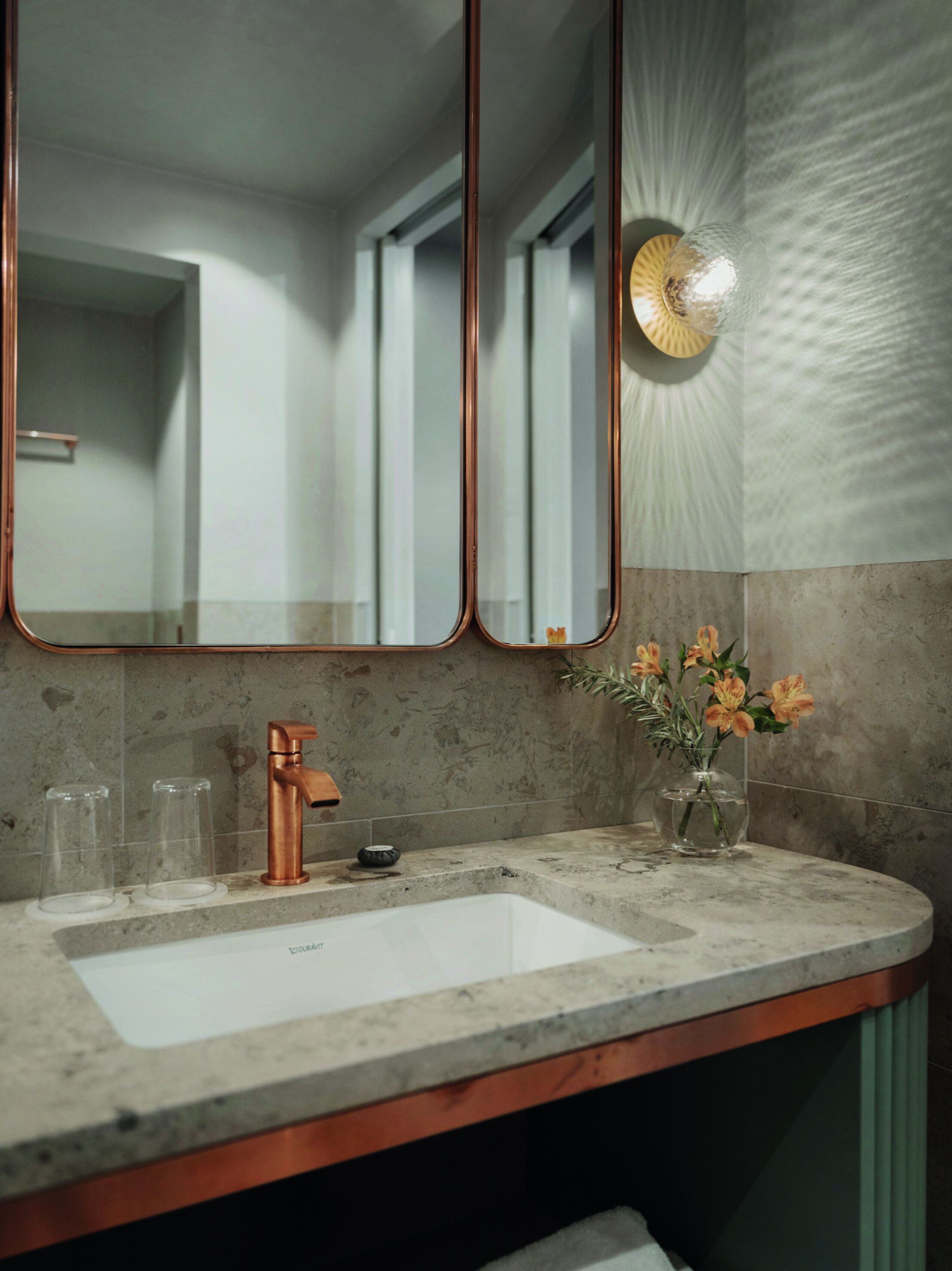 Un baño con un espejo grande, un grifo de cobre y un florero con flores en la encimera.