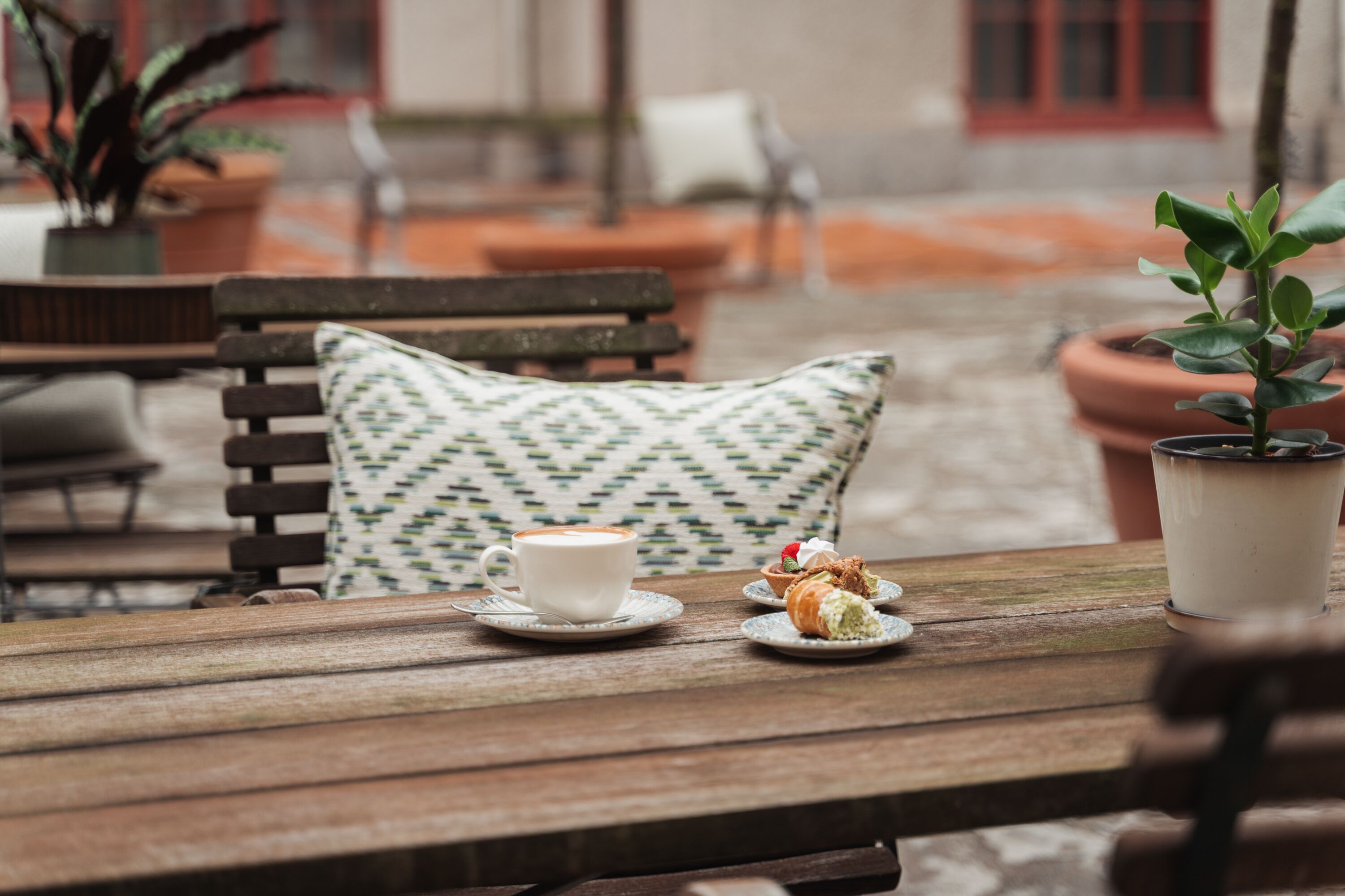 A wooden table with a white cup and a pastry, a patterned pillow, and a potted plant.