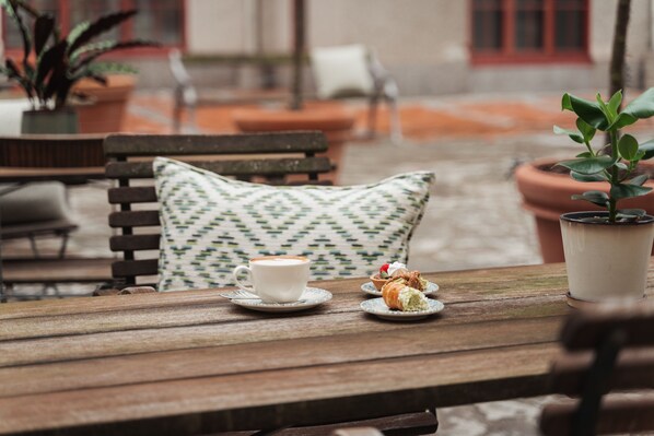 Una mesa de madera con una taza blanca y un pastel, un cojín estampado y una planta en maceta.