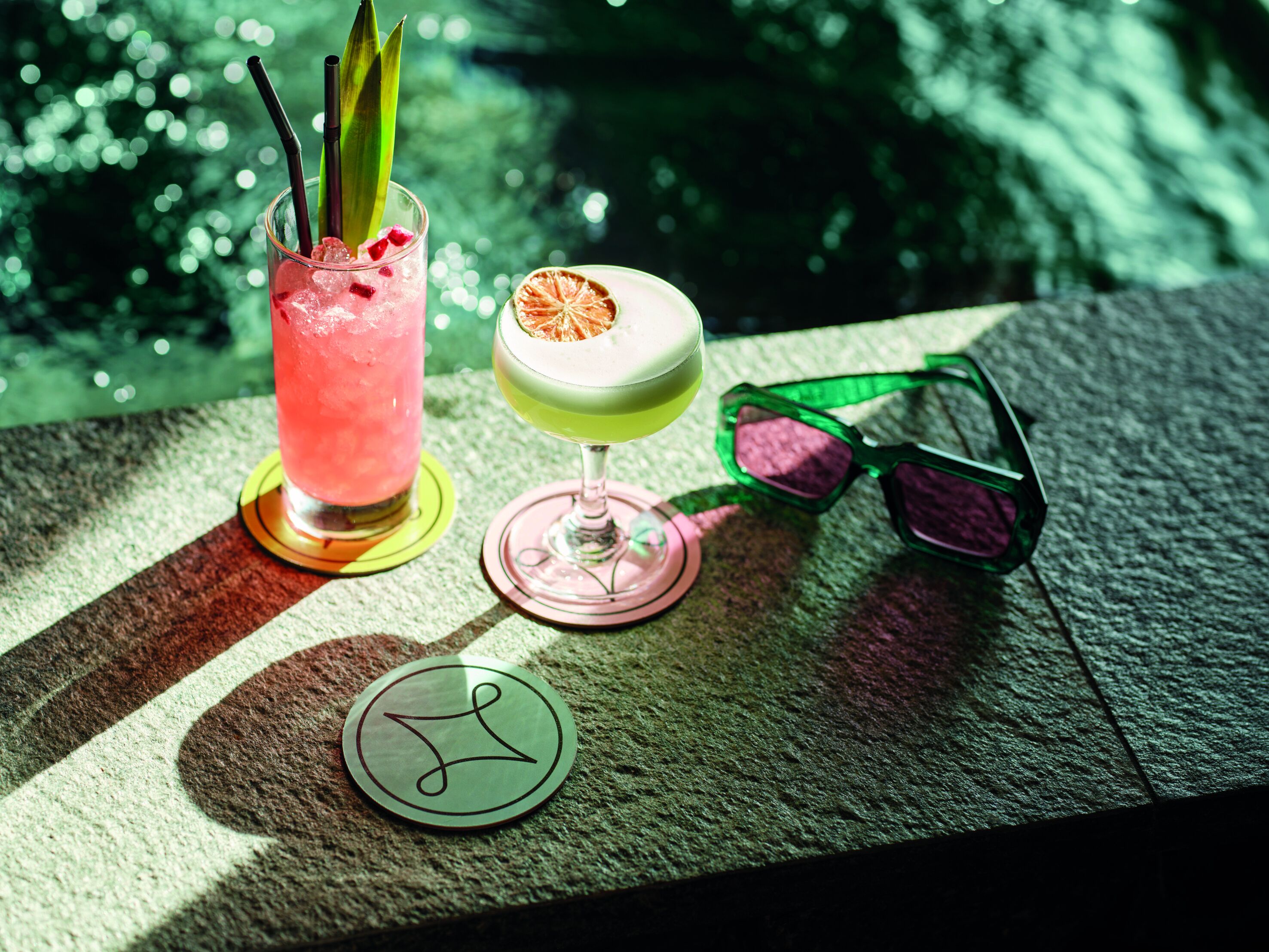 Two cocktails on a table with a pair of sunglasses and a coaster with a logo.