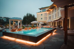 A rectangular pool with lounge chairs and umbrellas, surrounded by stone flooring and a building with multiple windows and balconies.