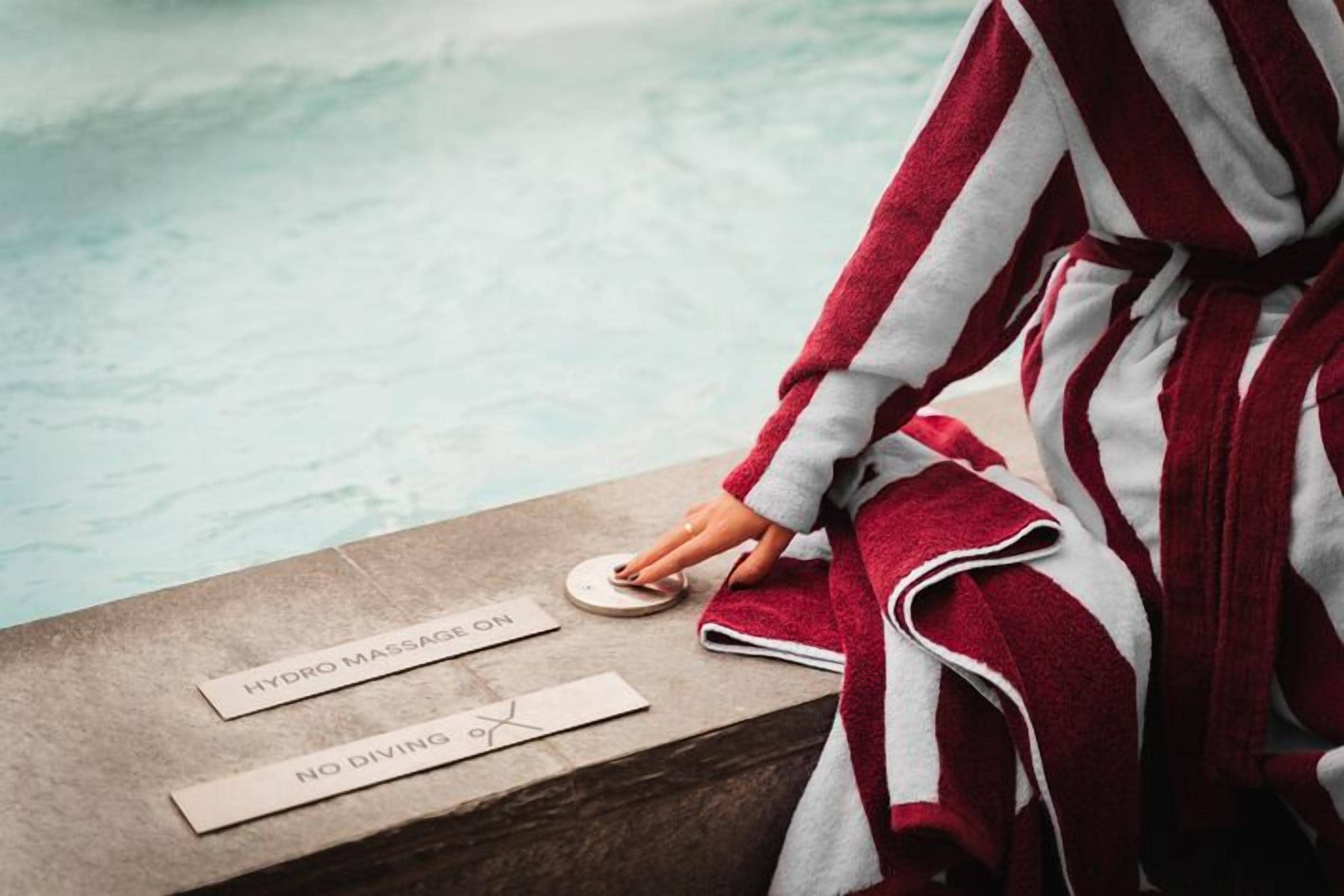 Une personne vêtue d’une robe rayée rouge et blanche se tient près d’une piscine, à côté de panneaux indiquant « HYDROMASSAGE EN FONCTION » et « INTERDIT DE PLONGER ».
