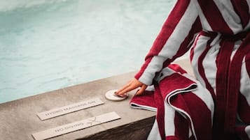 Une personne vêtue d’une robe rayée rouge et blanche se tient près d’une piscine, à côté de panneaux indiquant « HYDROMASSAGE EN FONCTION » et « INTERDIT DE PLONGER ».