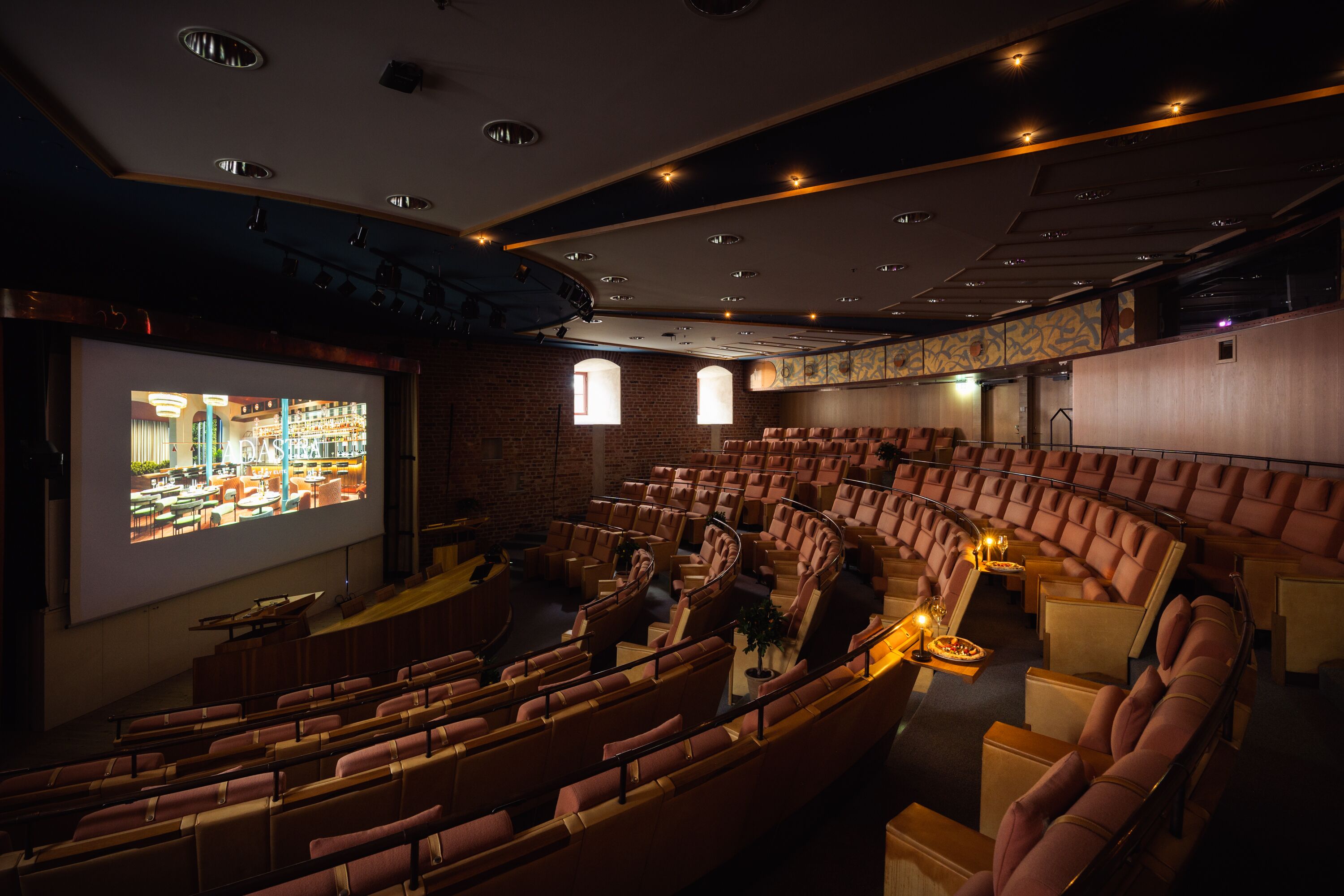 Un teatro con butacas escalonadas, una pantalla grande y una zona de bar visible a través de las ventanas.