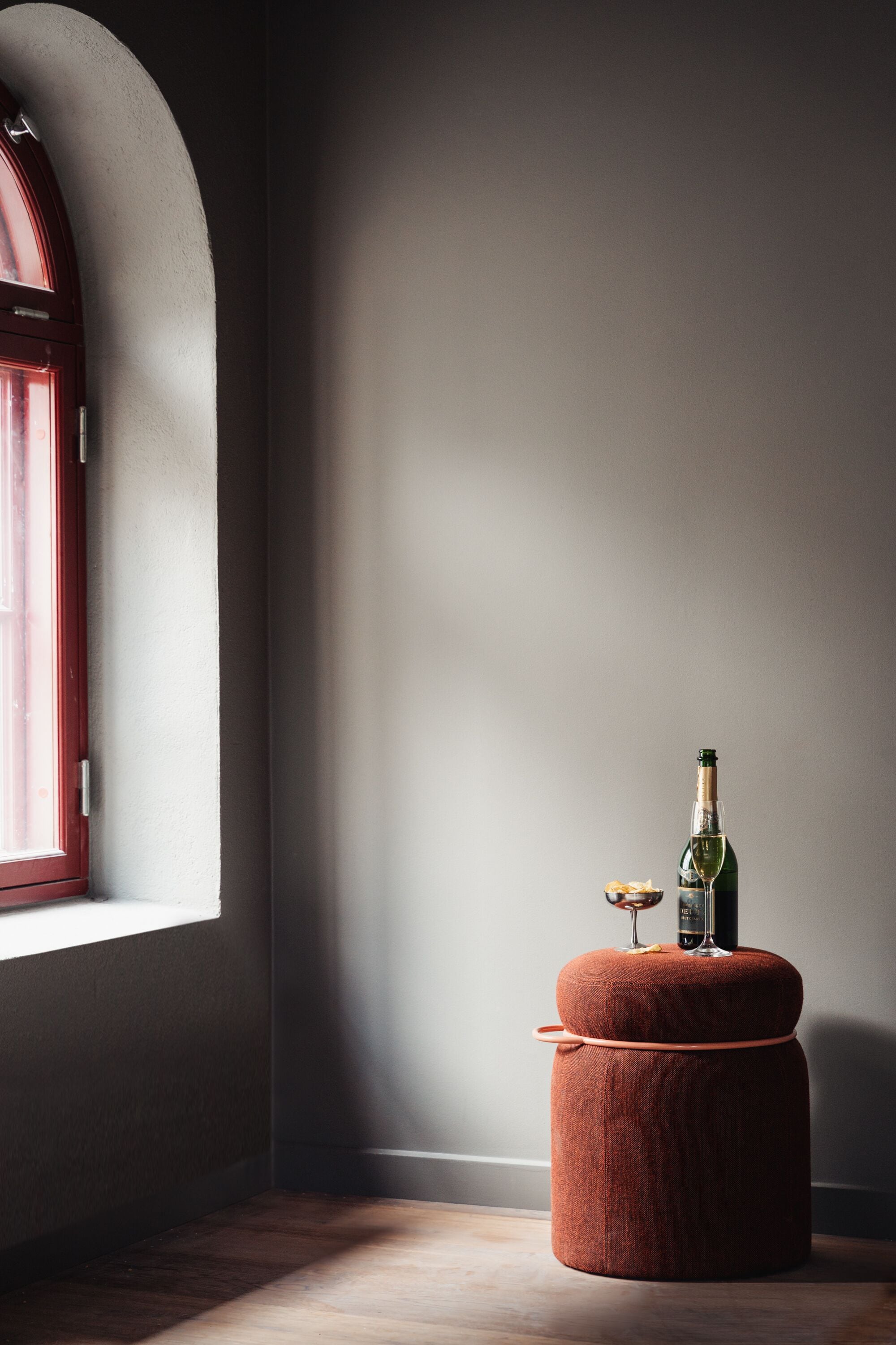 A room with a window, a red door, and a small ottoman with a bottle and glass.