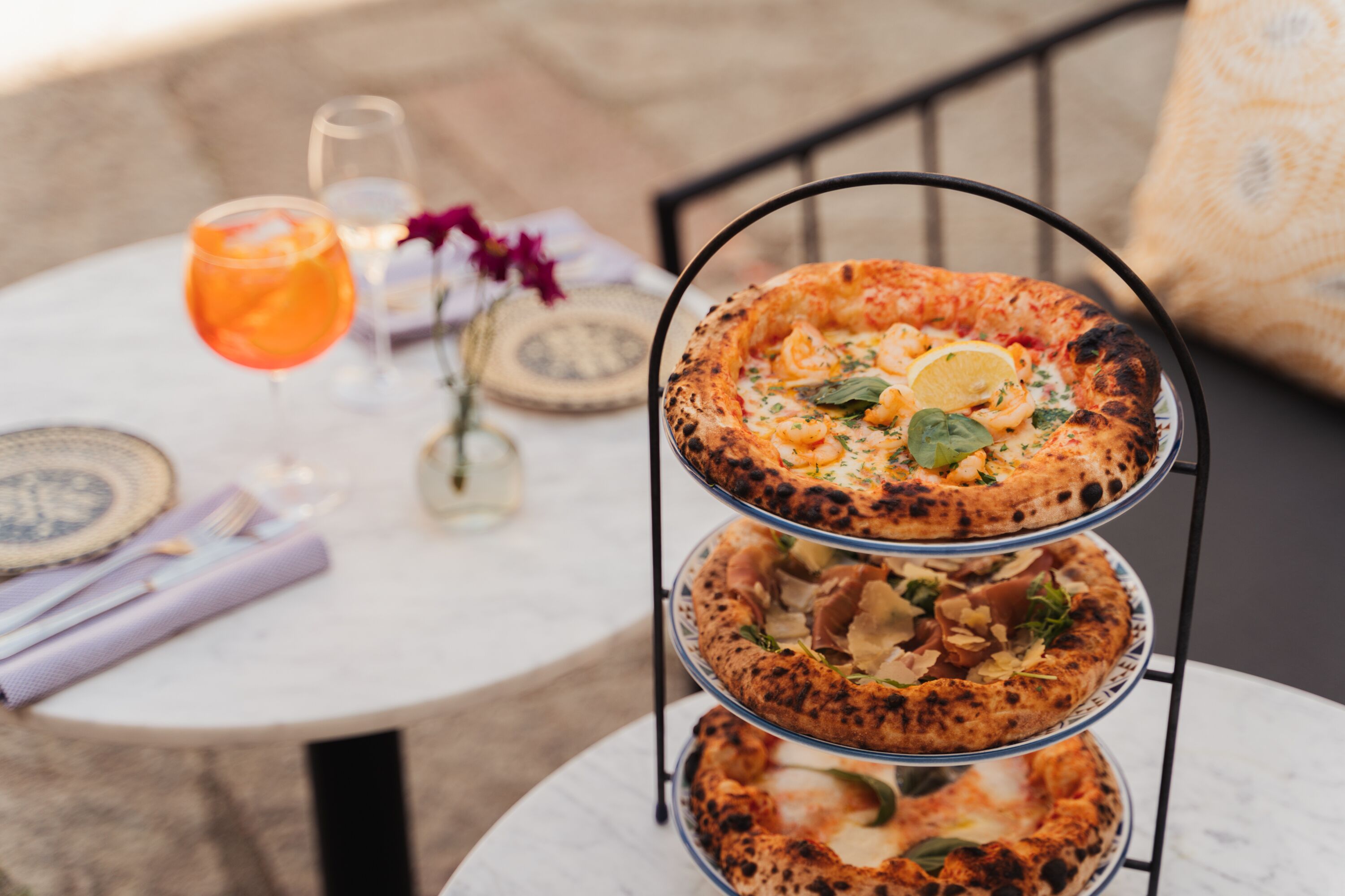Ett pizzaställ med tre våningar, med olika sorters pizzor, ett glas apelsinläsk och en liten vas med blommor på ett runt bord.