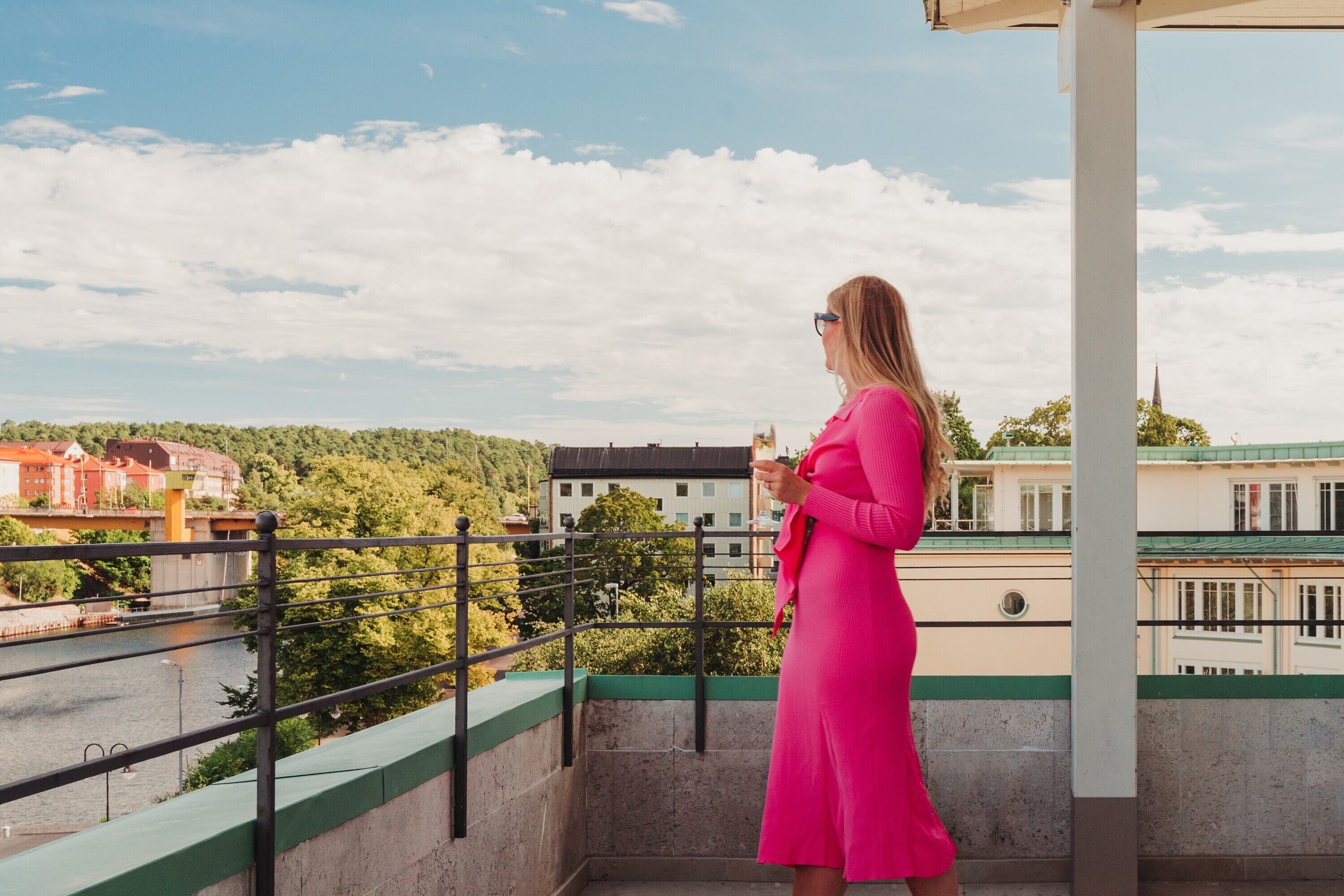 Una mujer con un vestido rosa parada en un balcón con vistas a un río y edificios.