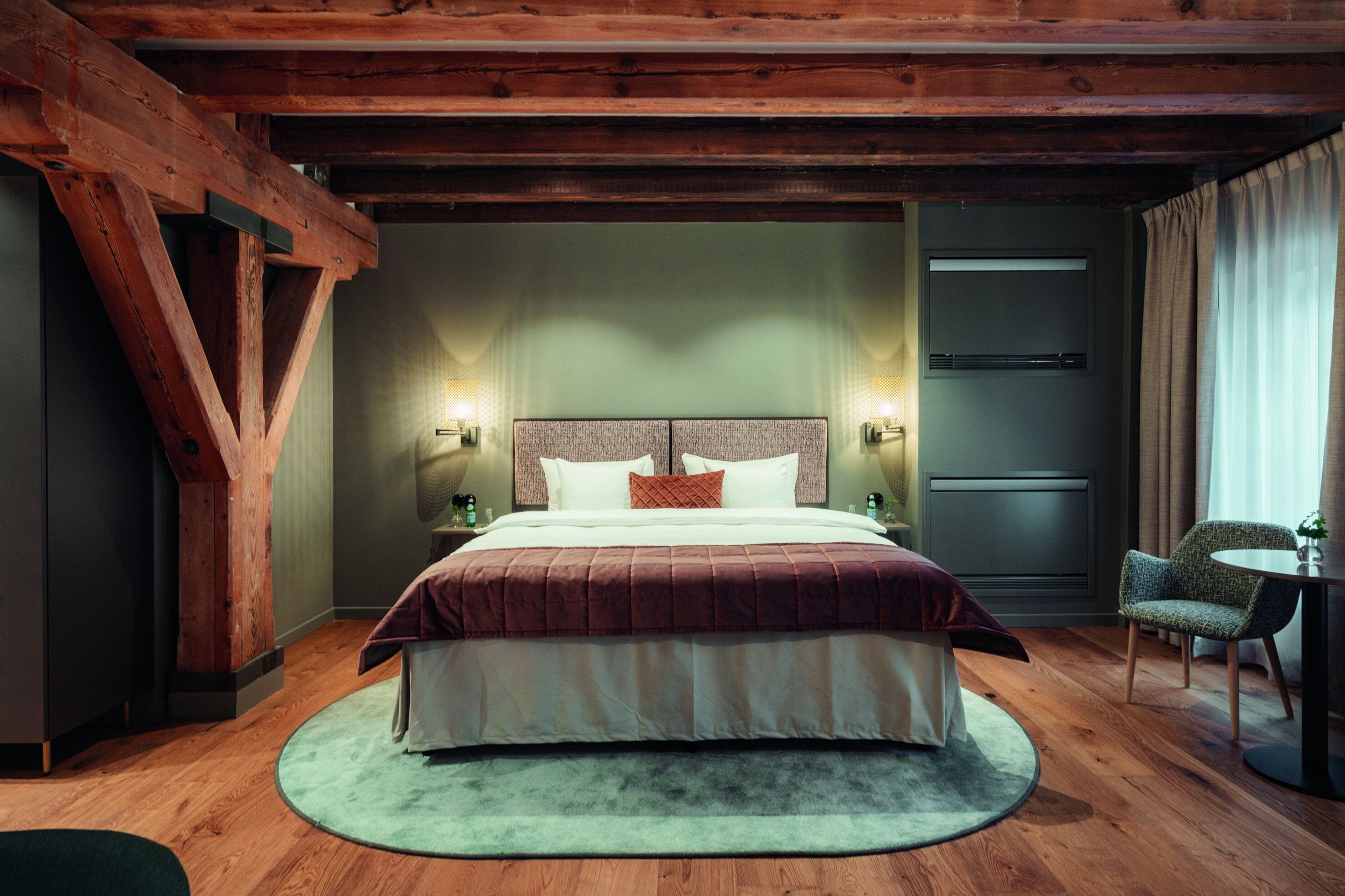 A hotel room with a large bed, wooden beams, a round rug, a small table, and a chair.