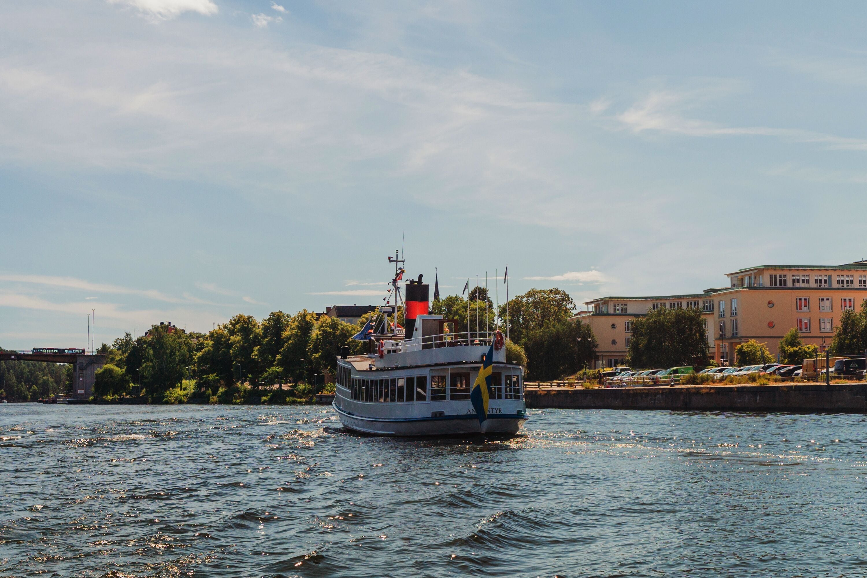 A riverboat with a flag on its mast, moving on a river with a cityscape in the background.
