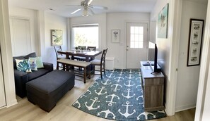 A living room with a dark sofa, a dining table with chairs, a TV on a wooden stand, and a patterned rug.