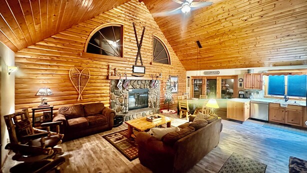 A log cabin interior with a fireplace, wooden furniture, a ceiling fan, and a kitchen area.