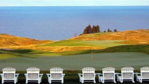A golf course with a view of the ocean, a clubhouse, and several white chairs in the foreground.