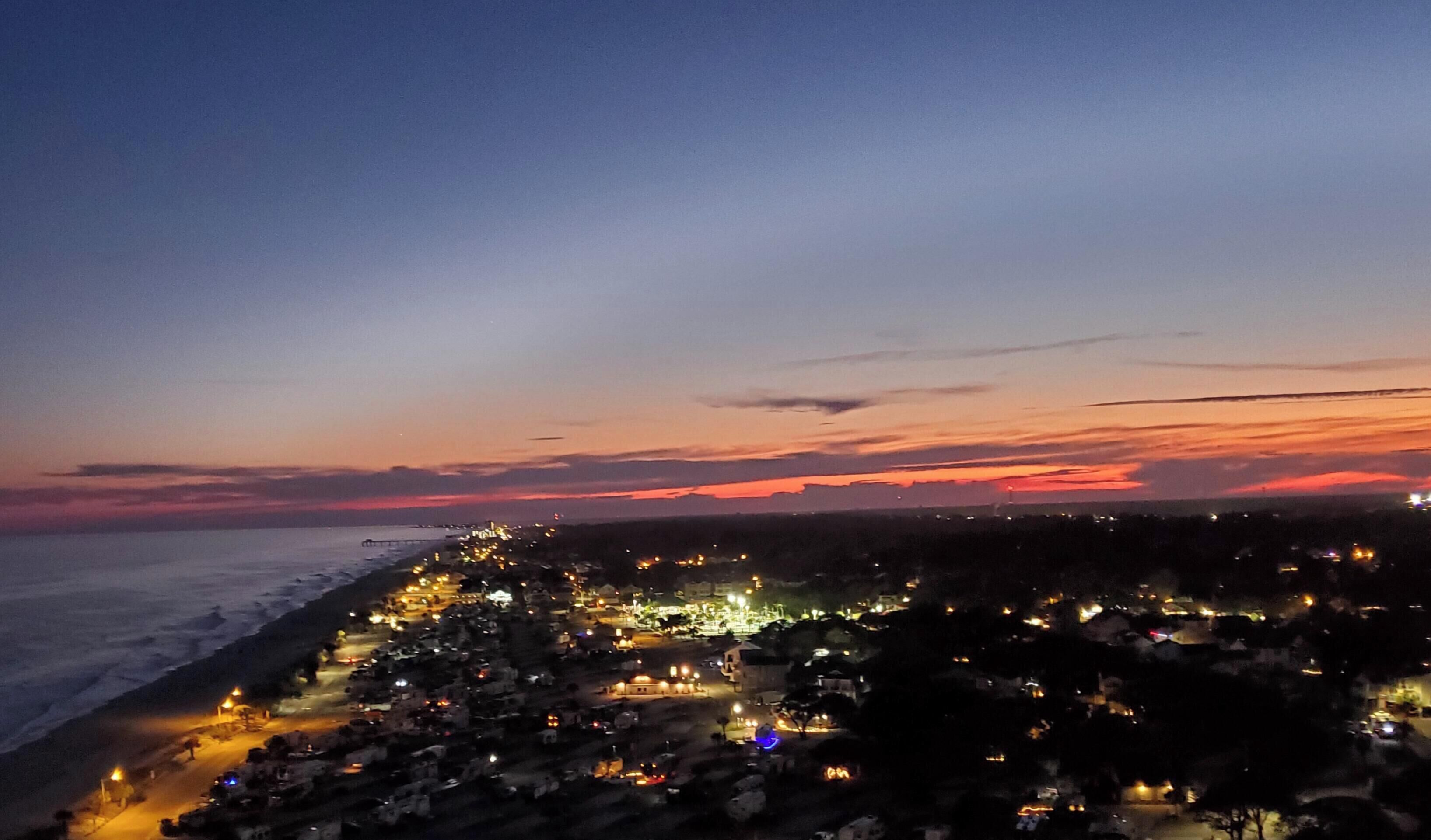 A coastal town at dusk with a view of the ocean and a long stretch of illuminated buildings along the shore.