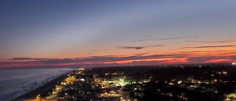A coastal town at dusk with a view of the ocean and a long stretch of illuminated buildings along the shore.