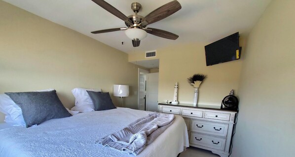 Quarto com cama, penteadeira, ventilador de teto e TV na parede.