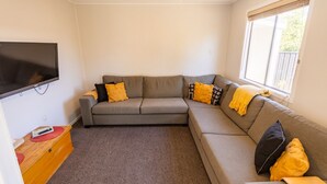 A living room with a grey sectional sofa, a flat-screen TV mounted on the wall, a wooden side table with a remote control, and a window with blinds.