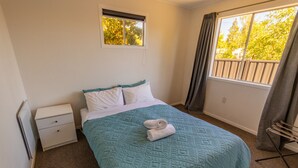 A single bed with a teal quilt, white pillows, and a folded towel on the bed. There is a small bedside table with a drawer, a window with a view of trees, and a curtain on the window.
